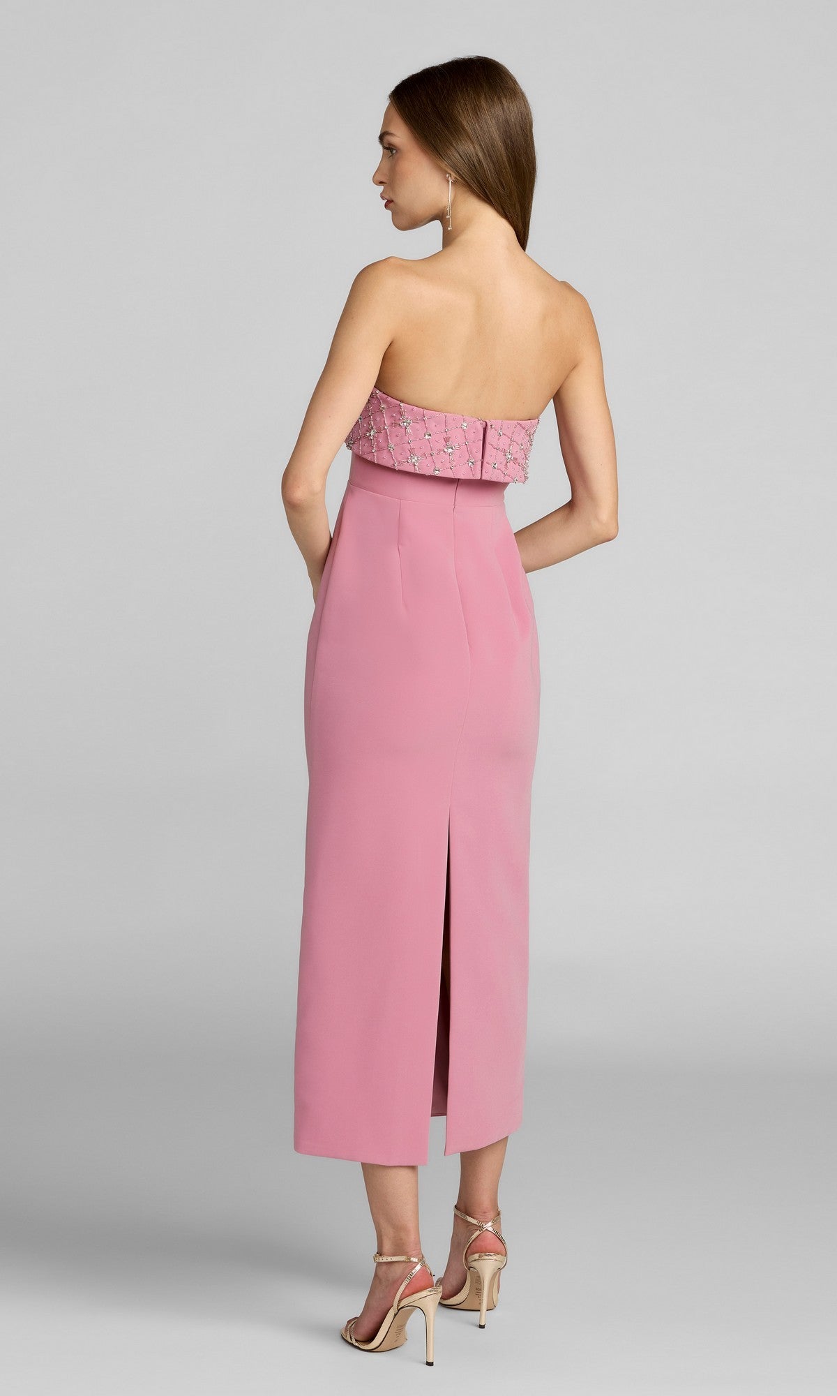 A woman faces away, wearing the Mac Duggal 11883 midi wedding guest dress in pink—strapless with rhinestone bodice embellishments and a back slit—paired with nude high-heeled sandals.